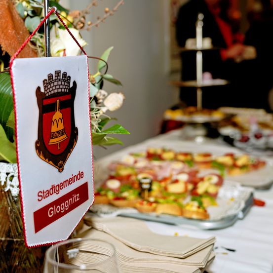 Ein Banner mit dem Text 'Stadtgemeinde Gloggnitz' hängt an einem Stand mit Blumen. Dahinter steht ein Tisch mit Essensplatten, und eine Person steht in der Nähe eines Dessert-Standes.