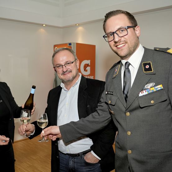 Drei Personen versammeln sich drinnen und lächeln. Ein Mann in Uniform stößt mit einem Mann in Anzug an, während eine Frau eine Flasche Wein hält. Im Hintergrund ist ein Großbuchstabe 'G' zu sehen.