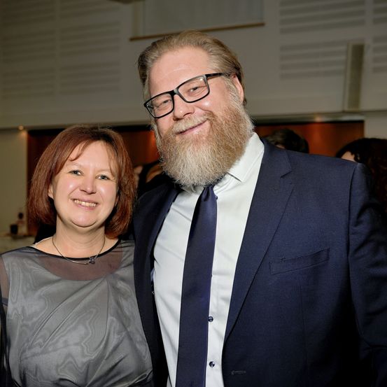 Ein Mann mit Bart in Anzug und Krawatte steht neben einer lächelnden Frau in einem grauen Kleid bei einer Veranstaltung.