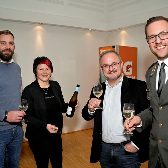Vier Personen in formeller Kleidung, lächelnd, halten Weingläser. Eine Frau mit roten Haaren auf der linken Seite, ein Mann mit Brille, ein uniformierter Mann und ein bärtiger Mann. Sie befinden sich in einem Raum mit einer weißen Wand und einem gelben Kasten mit dem Buchstaben G.