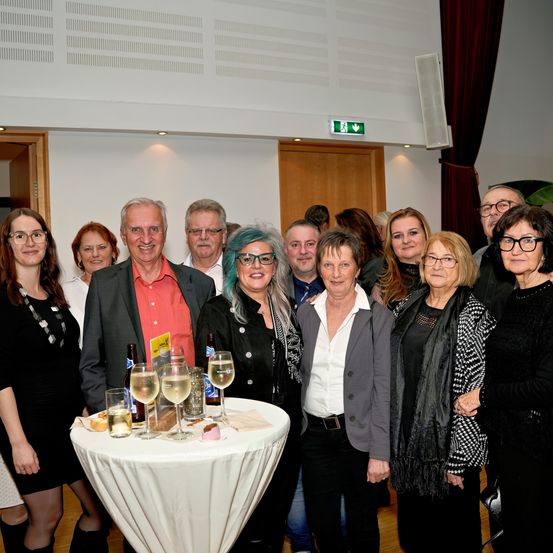 Eine Gruppe Erwachsener steht um einen Tisch mit Weinglasern, einem Kuchen und Flaschen. Sie sind festlich gekleidet. Hinter ihnen ist ein Raum mit einer weißen Wand und einer offenen Tür zu sehen.