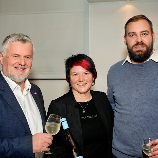 Drei Personen stehen zusammen, lächeln und halten Weingläser. Der Mann auf der linken Seite trägt einen Anzug, die Frau in der Mitte hat rote Haare und eine Halskette, und der Mann auf der rechten Seite trägt einen grauen Pullover. Sie stehen vor einer weißen Wand.