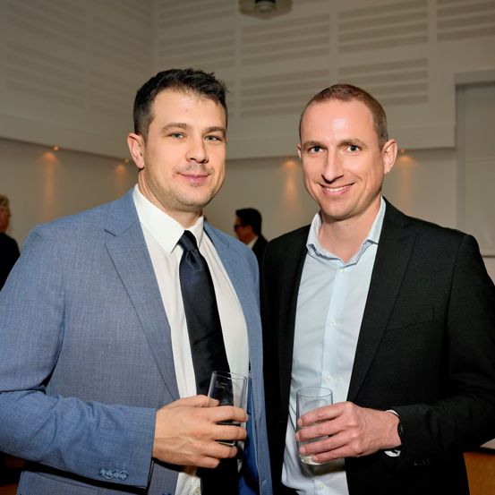 Zwei Männer in Anzügen, lächelnd, stehen bei einer Veranstaltung zusammen. Sie halten Gläser mit Getränken in den Händen.