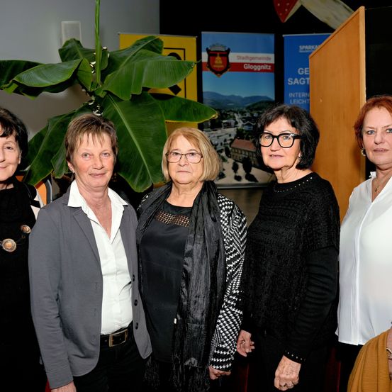 Fünf Frauen stehen zusammen vor einer Kulisse mit einer großen Pflanze und Schildern. Sie lächeln und scheinen sich auf einer Veranstaltung zu befinden.