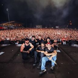 Eine Rockband posiert für ein Foto auf der Bühne bei einem nächtlichen Konzert, umgeben von einem begeisterten Publikum.
