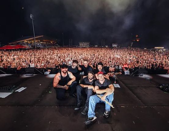 Eine Rockband posiert für ein Foto auf der Bühne bei einem nächtlichen Konzert, umgeben von einem begeisterten Publikum.