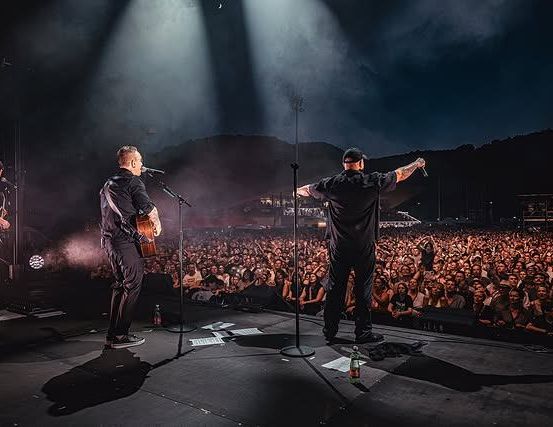 Eine Band spielt bei Nacht auf der Bühne vor einem großen Publikum. Ein Mitglied hält eine Gitarre, ein anderes ein Mikrofon.