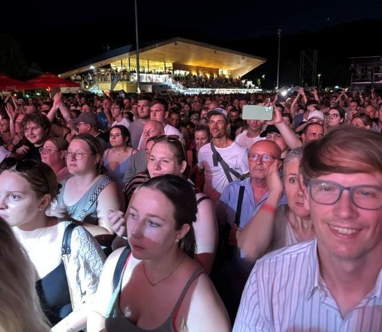 Eine große Menschenmenge versammelt sich bei Nacht für eine Veranstaltung im Freien. Einige Menschen halten Telefone, um den Moment festzuhalten. Ein Mann lächelt im Vordergrund.
