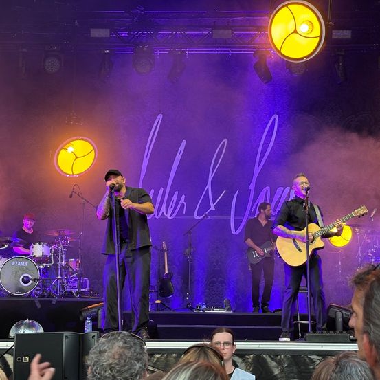 Eine Band tritt auf der Bühne mit lila und gelben Lichtern auf. Der Leadsänger steht links mit einem Mikrofon. Ein Gitarrist spielt rechts. Der Schlagzeuger steht hinter einem Schlagzeug. Das Publikum sieht von unten zu.