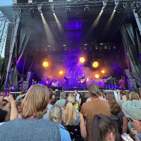 Ein Live-Konzert findet auf der Bühne statt, mit einem großen Publikum. Die Band spielt Gitarren und singt auf der Bühne mit heller Beleuchtung. Das Publikum sieht zu und genießt die Aufführung.