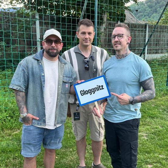 Drei Männer stehen auf einer Rasenfläche. Ein Mann hält ein Schild mit der Aufschrift 'Gloggnitz'. Hinter ihnen ist ein grünes Netz und Bäume.