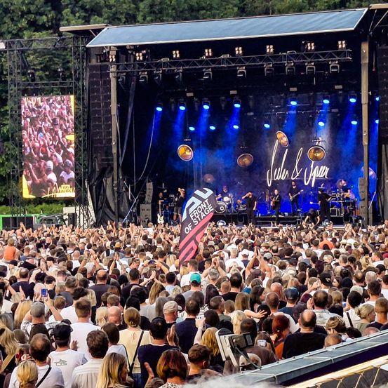 Ein Publikum versammelt sich bei einem Konzert, mit einer Bühne, die in blaues Licht getaucht ist und einem großen Bildschirm hinter der Band. Das Publikum ist energiegeladen und hält Schilder hoch und hebt die Hände.