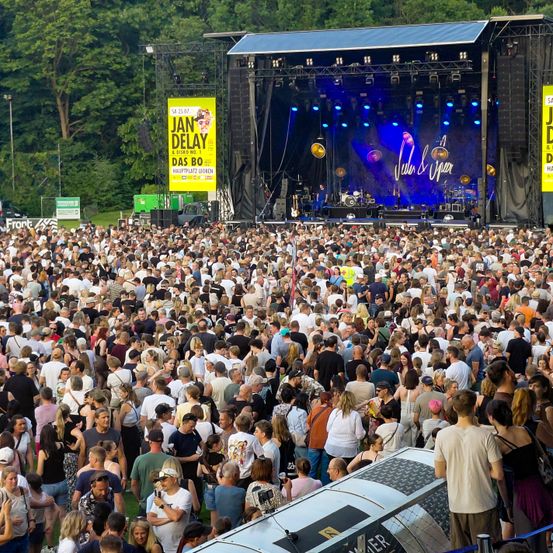 Eine große Menschenmenge versammelt sich für ein Open-Air-Musikfestival. Die Bühne ist mit einem Banner für Jan Delay ausgestattet. Künstler sind auf der Bühne unter einer blauen Lichtinstallation.
