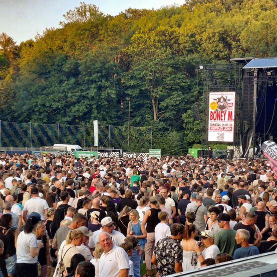 Eine große Menschenmenge hat sich für ein Konzert versammelt, steht vor einer Bühne mit einem Plakat für Boney M. Im Hintergrund sind Bäume und ein klarer Himmel zu sehen.