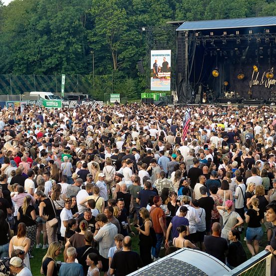 Eine große Menschenmenge versammelt sich bei einem Open-Air-Konzert. Die Bühne ist mit einem Banner und Lichtern ausgestattet. Das Publikum ist vielfältig, viele tragen Hüte, Brillen und tragen Taschen. Einige stehen, andere sitzen auf dem Gras. Die Bäume im Hintergrund deuten auf eine natürliche Umgebung hin.