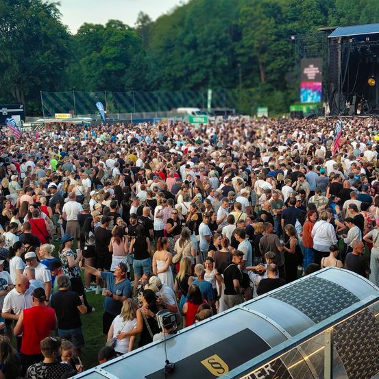 Eine große Menschenmenge versammelt sich im Freien für eine Musikveranstaltung, steht und mischt sich auf Rasenflächen. Die Bühne ist mit einer großen Leinwand und Beleuchtungsequipment ausgestattet.