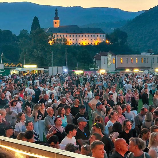 Eine Menschenmenge versammelt sich am Abend bei einem Festival. Ein großes Gebäude ist im Hintergrund zu sehen.