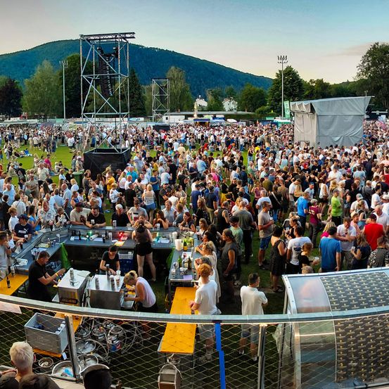 Eine große Menschenmenge versammelt sich bei einer Veranstaltung im Freien. Sie unterhalten sich und trinken in der Nähe von Essensständen und einer Bühne. Berge sind im Hintergrund zu sehen.
