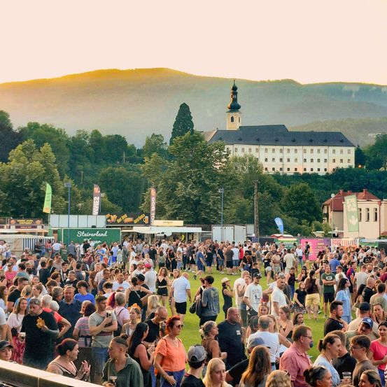 Eine große Menschenmenge versammelt sich im Freien mit Bergen im Hintergrund. In der Mitte ein weißes Gebäude mit Turm und vielen Fenstern. Es sind Essenswagen und Schilder zu sehen.