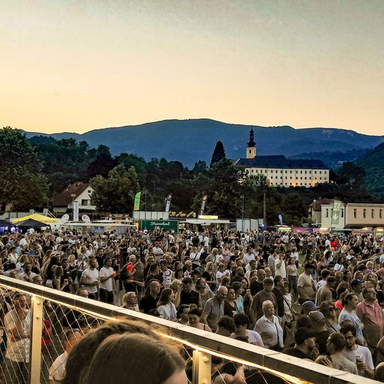 Eine große Menschenmenge versammelt sich für eine Veranstaltung im Freien. Bäume und Berge bilden die Kulisse. Die Umgebung ist lebendig mit vielen Menschen und einem Gefühl der Gemeinschaft.