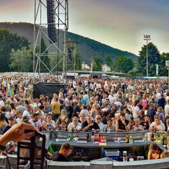 Eine große Menschenmenge versammelt sich für eine Outdoor-Musikveranstaltung mit Bergen im Hintergrund. Es gibt Essensstände, eine Bar und hohe Lautsprecher.