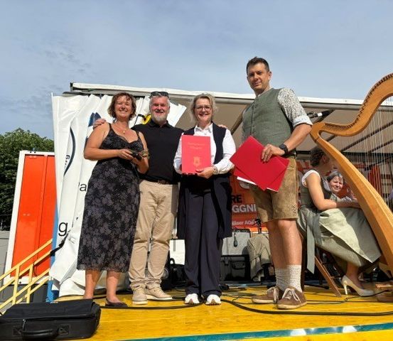 Vier Personen stehen lächelnd auf einer Bühne und halten rote Dokumente in der Hand. Ein Harfe steht hinter ihnen. Eine Frau sitzt auf einem Stuhl mit einer Harfe. Bäume und ein weißes Banner sind im Hintergrund.