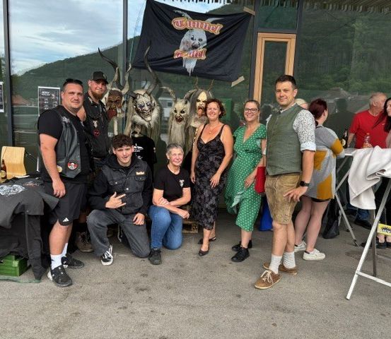 Eine Gruppe von Menschen posiert für ein Foto im Freien. Einige knien, andere stehen. Einige tragen Alltagskleidung, andere Kostüme. Ein Banner und Plakate hängen an der Wand.