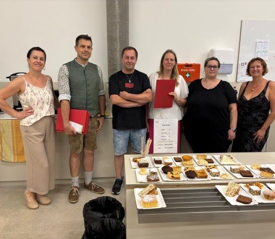 Eine Gruppe von Menschen, darunter Männer und Frauen, steht hinter einem Tisch mit verschiedenen Desserts. Sie tragen alle Freizeitkleidung und lächeln. Dahinter befindet sich eine Tafel und ein Schrank mit einem gelben Handtuch.