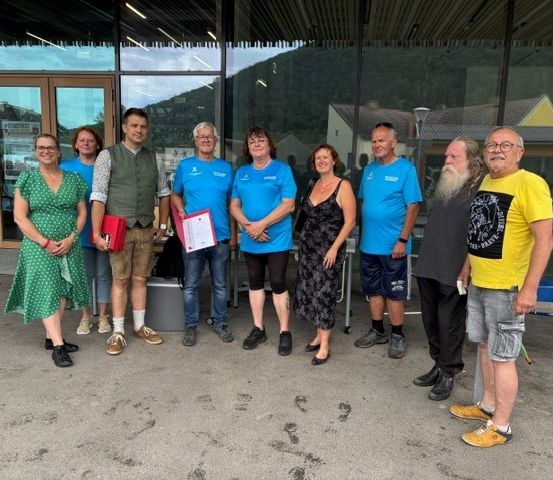 Eine Gruppe von Menschen in Freizeitkleidung steht vor einem Gebäude und hält Papiere in der Hand. Sie scheinen für ein Foto zu posieren.