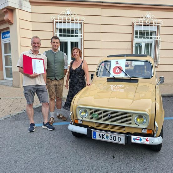 Drei Personen stehen neben einem beigefarbenen Renault-Auto mit einem Nummernschild 6 auf der Motorhaube. Ein Mann hält eine rote Mappe und ein Zertifikat. Das Auto hat das Kennzeichen NK 30 D.