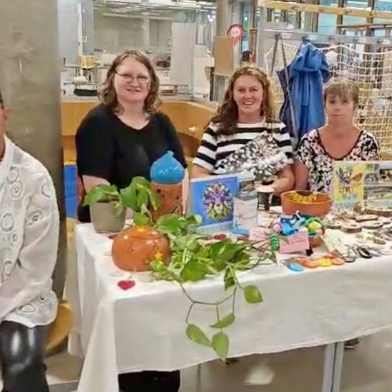 Vier Frauen stehen lächelnd hinter einem Tisch mit verschiedenen Gegenständen. Der Tisch ist mit Pflanzen, einer Kürbis und Büchern geschmückt. Im Hintergrund befinden sich ein Aufbewahrungsbox und ein Netz.