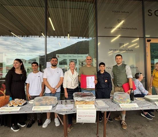 Gruppe von Personen, darunter Erwachsene und Kinder, steht vor einem Tisch mit verschiedenen Lebensmitteln in einer Außenumgebung in der Nähe eines Schulgebäudes.