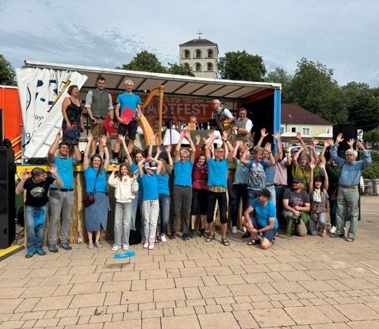 Eine Gruppe von Menschen in blauen Shirts steht vor einem Fahrzeug mit einer Fahne. Das Fahrzeug ist vor einem Haus mit einem Turm geparkt.