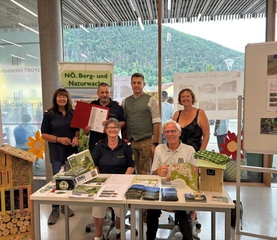 Eine Gruppe von Menschen steht an einem Tisch mit verschiedenen Materialien, lächelt und posiert für ein Foto. Der Tisch hat Broschüren und ein Bienenstockmodell. Der Hintergrund besteht aus einer Glaswand mit Naturansichten.