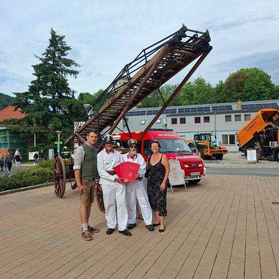 Vier Personen stehen vor einem historischen Feuerwehrauto, zwei von ihnen in traditioneller Uniform, einer hält ein rotes Buch, eine Frau in einem schwarzen Kleid, und verschiedene Fahrzeuge und Bäume im Hintergrund.