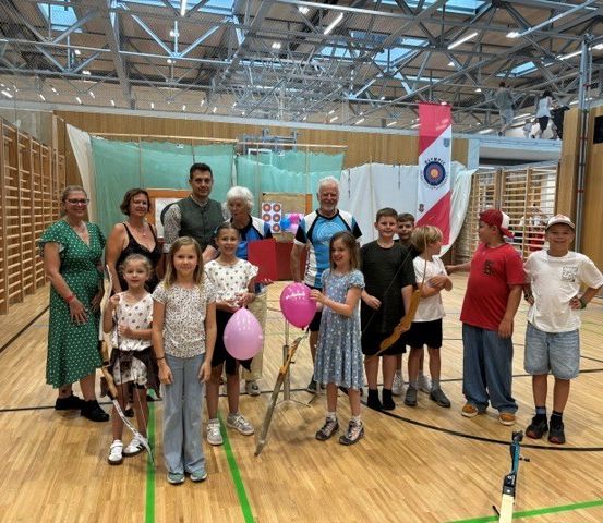 Eine Gruppe von Menschen, einschließlich Erwachsener und Kindern, versammelt sich in einer Sporthalle. Sie posieren für ein Foto mit Luftballons und Bögen.