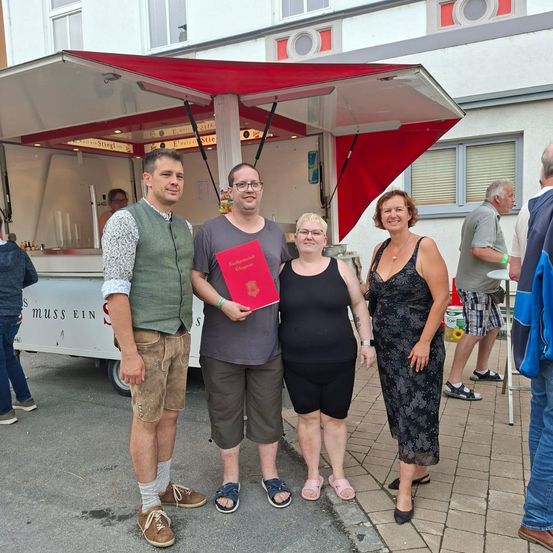 Eine Gruppe von Menschen steht vor einem Essenswagen. Drei Menschen halten ein rotes Buch, und andere stehen hinter ihnen. Ein Mann schaut in die Kamera, und ein anderer Mann lächelt. Eine Frau trägt eine Halskette und ein Armband.