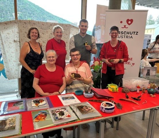 Eine Gruppe von Menschen, alle in Rot gekleidet, steht hinter einem Tisch. Sie lächeln für ein Foto. Der Tisch hat verschiedene Gegenstände darauf, einschließlich Bücher, einen Korb und eine Blume. Dahinter steht ein großes Banner mit einem Herzsymbol und Text in einer Fremdsprache.