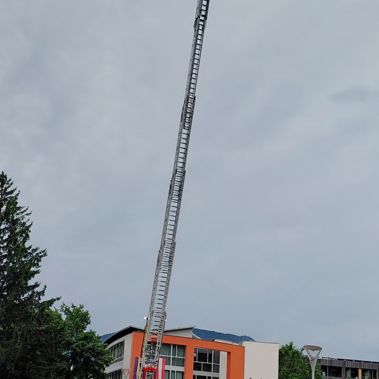 Eine hohe Feuerwehrleiter steht gegen einen bewölkten Himmel in der Nähe eines Gebäudes mit orangefarbenen Akzenten. Bäume umgeben das Gebiet, eine Straßenlaterne ist rechts sichtbar.