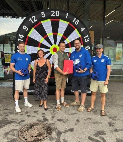 Fünf Personen stehen vor einem großen Dartboard, halten Fußballbälle und eine rote Mappe. Sie scheinen einen Sieg oder eine Errungenschaft zu feiern.