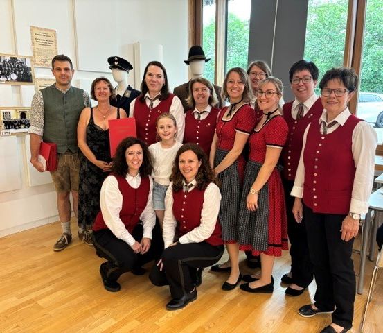 Eine Gruppe von Menschen in traditioneller bayerischer Kleidung posiert für ein Foto in einem Raum. Mannequins und Poster schmücken die Wände.