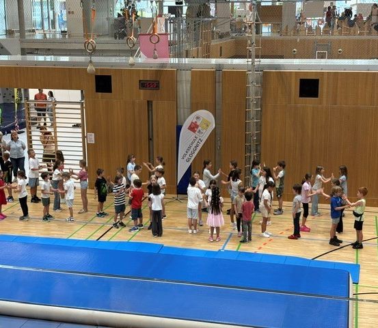 In einer Turnhalle versammelt sich eine große Gruppe von Kindern, die verschiedene Kleidungsstücke und Schuhe tragen, spielen und interagieren. Ein großer blauer Teppich liegt auf dem Boden, und es sind Erwachsene in der Nähe.
