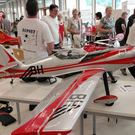 Eine Gruppe von Menschen in einem Raum, einige stehen um einen Tisch herum, auf dem ein rotes und weißes Modellflugzeug steht. Das Flugzeug hat die Buchstaben BH auf sich.