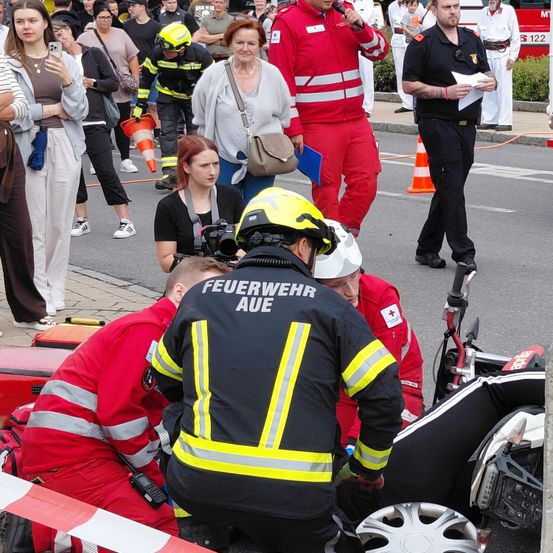 Einsatzkräfte, einschließlich Feuerwehrleute, kümmern sich um eine verletzte Person auf einer Straße. Schaulustige versammeln sich, und andere gehen oder stehen in der Nähe. Ein Feuerwehrwagen ist im Hintergrund zu sehen.