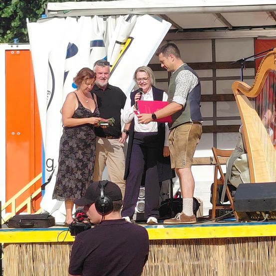 Eine Gruppe von Menschen steht auf der Bühne, ein Mann hält ein Mikrofon und eine Mappe und eine Frau schaut sich ein Blatt Papier an. Dahinter stehen eine Harfe und ein Lautsprecher.