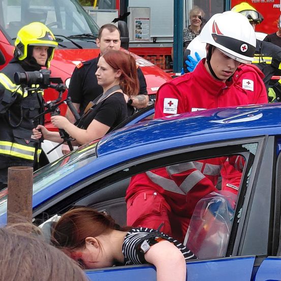 Rettungskräfte kümmern sich um eine verletzte Frau in einem blauen Auto. Ein Feuerwehrmann filmt mit einer Kamera. Eine Frau in einer roten Uniform und einem Helm hilft der Verletzten.