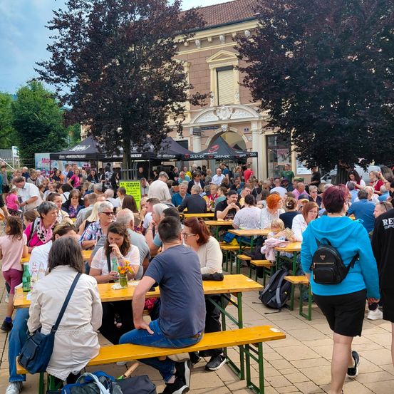 Eine große Menschenmenge ist draußen versammelt, sitzt an Picknicktischen, einige mit Getränken. Bäume und ein Gebäude mit Vordächern im Hintergrund. Einige Leute stehen und gehen herum.