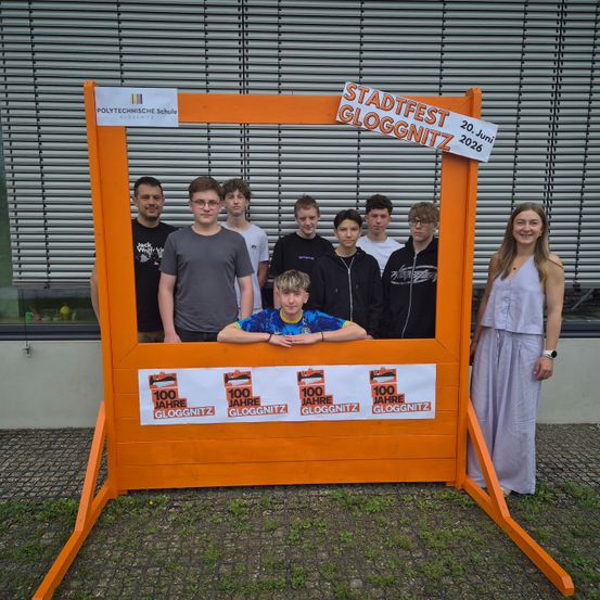 Eine Gruppe von Schülern und eine Frau posieren hinter einem Fotostand für das Gloggnitz Stadtfest. Der Stand ist orange mit einem weißen Schild und steht '100 Jahre Gloggnitz'. Das Festival ist am 20. Juni 2026.