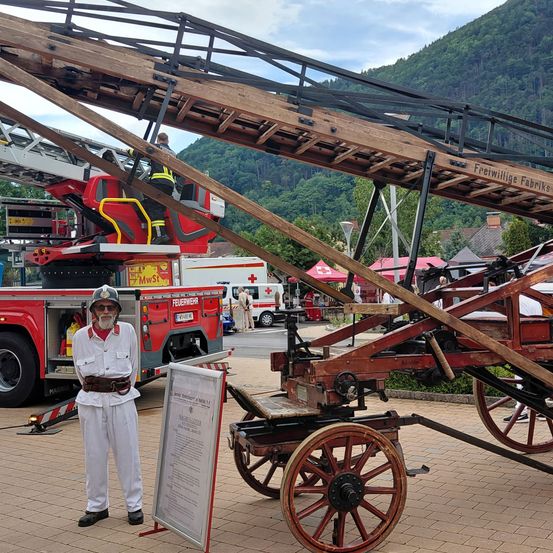 Ein Mann steht neben einem alten Feuerwehrwagen auf einem Ziegelboden. Hinter ihm ist ein Feuerwehrwagen mit ausgefahrener Leiter geparkt, und eine Person steht auf der Leiter. Der Hintergrund zeigt einen Berg und Bäume.