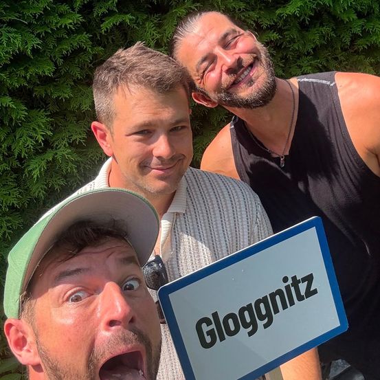 Drei Männer stehen vor grünen Büschen und posieren für ein Foto. Einer trägt ein gestreiftes Hemd, der andere ein schwarzes Tanktop. Sie alle lächeln. Ein Schild mit der Aufschrift Glogginitz wird von einem der Männer gehalten.